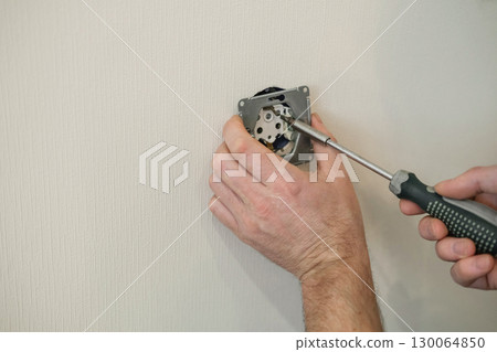 Close-up of hands screwing socket with a screwdriver against light wall. Home renovation, electrical work, DIY projects and maintenance 130064850