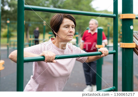 elderly woman doing exercises on a horizontal bar at an outdoor sports ground 130065257