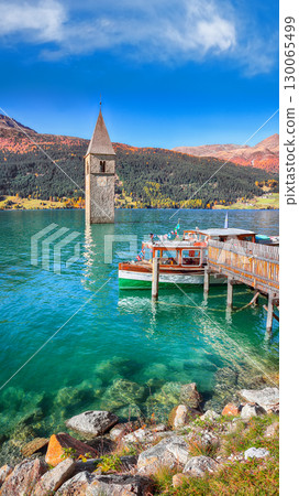 Astonishing autumn lake landscape with a ship near the pier and submerged bell tower in lake Resia or Reschensee. Astonishing autumn lake landscape with a ship near the pier and submerged bell tower in lake Resia or Reschensee. 130065499