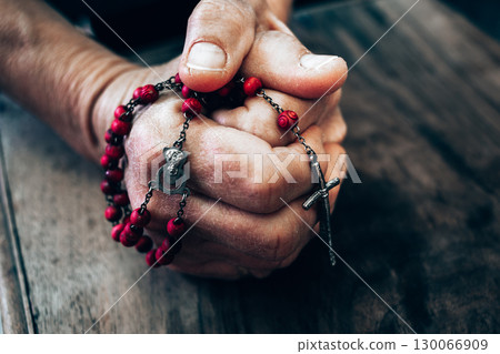 Weathered hands clutching crimson rosary beads in contemplative prayer on rustic wooden surface Weathered hands clutching crimson rosary beads in contemplative prayer on rustic wooden surface 130066909