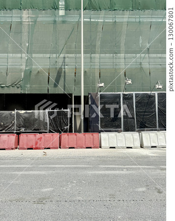 Street view of construction barriers in red and white, green safety mesh, black wrapped panels, and scaffolding in background, urban infrastructure and renovation 130067801