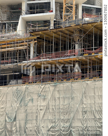 Modern building under construction with visible scaffolding, yellow framework, concrete columns, and protective green mesh netting, active urban development 130067802