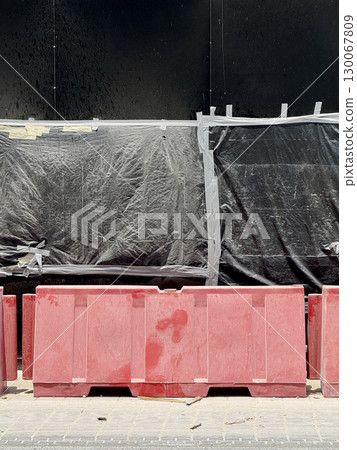 Close-up of red plastic construction barriers in front of black-wrapped panels, city pavement and renovation supplies, emphasizing urban infrastructure 130067809