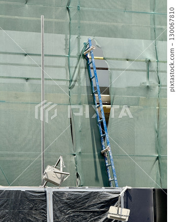 Detail of green mesh safety netting, blue ladder, floodlights, and part of building under renovation, illustrating city construction and equipment setup 130067810