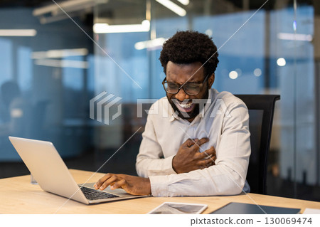 A man is sitting at a desk, experiencing chest pain while using a laptop in an office setting. This evokes health concerns or emergency awareness of heart-related issues in professional environments 130069474
