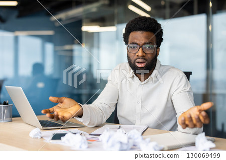 A professional in a modern office looks concerned while gesturing, papers scattered on the desk, portraying workplace stress. The setting includes a laptop, pens, and a professional attire. 130069499