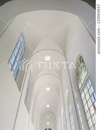 High-angle view of a stark white, modern hallway with a high vaulted ceiling, illuminated by large windows and a series of minimalist globe pendant lights hanging in a row 130069657