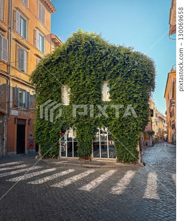 ROME, ITALY - AUGUST 01 2025 A building completely covered in thick green ivy stands at a cobblestone intersection. Charming Italian streets view 130069658