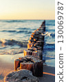 Wooden breakwater stretches into blue sea under soft evening light. Gentle waves touch sandy shore while sun reflects warmly on weathered timber posts and shimmering water 130069787