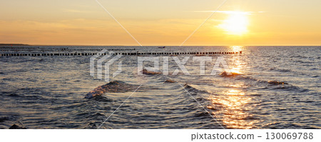Scenic warm sea sunset view golden sunset over Baltic Sea reflects across rippling waves, creating a glowing path of light. Wooden breakwaters stretch into horizon calm rhythm to tranquil seascape 130069788