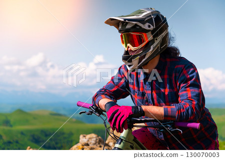 Portrait of a young man in a protective helmet and gloves, in bright clothes, sitting on a downhill bike and looking into the distance. Extreme rest in the mountains Portrait of a young man in a protective helmet and gloves, in bright clothes, sitting on a downhill bike and looking into the distance. Extreme rest in the mountains 130070003