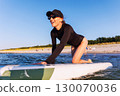 Smiling woman enjoys surfing on Baltic Sea waves, lying on board with splashing water around. Bright sunlight and her laughter reflect pure joy, freedom and vibrant summer adventure 130070036