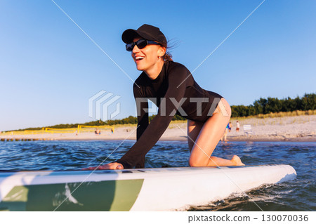 Smiling woman enjoys surfing on Baltic Sea waves, lying on board with splashing water around. Bright sunlight and her laughter reflect pure joy, freedom and vibrant summer adventure 130070036