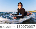 Smiling woman enjoys surfing on Baltic Sea waves, lying on board with splashing water around. Bright sunlight and her laughter reflect pure joy, freedom and vibrant summer adventure 130070037