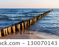 Wooden breakwater stretches into blue sea under soft evening light. Gentle waves touch sandy shore while sun reflects warmly on weathered timber posts and shimmering water 130070143