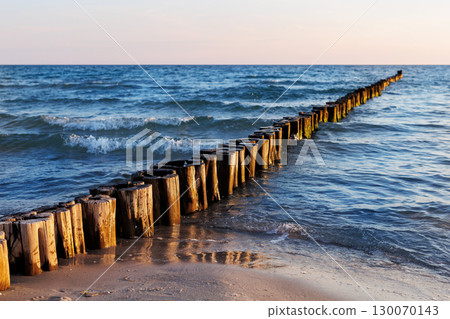 Wooden breakwater stretches into blue sea under soft evening light. Gentle waves touch sandy shore while sun reflects warmly on weathered timber posts and shimmering water 130070143