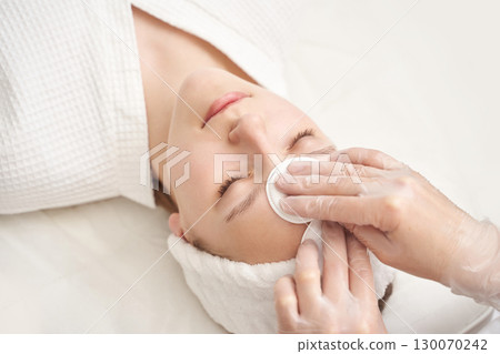 Young caucasian female receiving facial treatment with cotton pad in spa setting 130070242
