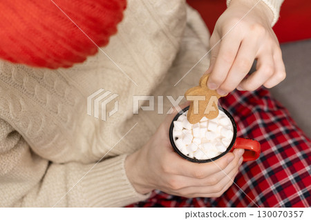 Young man in red plaid pajamas holding mug of hot cocoa topped with marshmallows and a gingerbread cookie. Festive atmosphere with warm lights and red beanie hat enhancing coziness. Xmas holiday. Home Young man in red plaid pajamas holding mug of hot cocoa topped with marshmallows and a gingerbread cookie. Festive atmosphere with warm lights and red beanie hat enhancing coziness. Xmas holiday. Home 130070357
