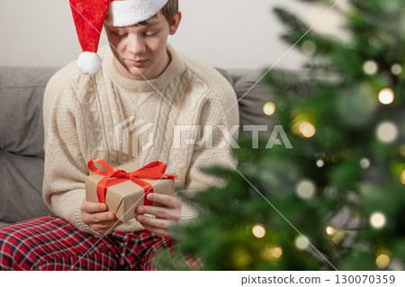 Sad young boy wearing Santa hat, white sweater and pajamas sits on sofa holding wrapped red gift. Cozy winter interior with Christmas tree and festive atmosphere. 130070359