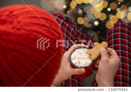 Young man sitting in red plaid pajamas holding mug of hot cocoa topped with marshmallows and gingerbread cookie. Christmas festive atmosphere with warm lights at cozy home interior. Xmas holiday. 130070361