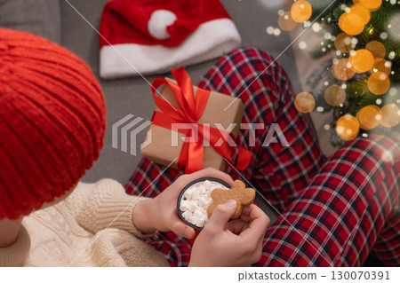 Young man in red plaid pajamas holding mug of hot cocoa topped with marshmallows and gingerbread cookie. Christmas festive atmosphere with warm lights at cozy home interior. Xmas holiday. 130070391
