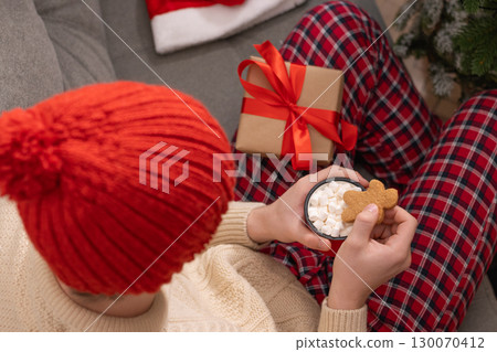 Young man in red plaid pajamas holding mug of hot cocoa topped with marshmallows and a gingerbread cookie. Festive atmosphere with warm lights and red beanie hat enhancing coziness. Xmas holiday. Home Young man in red plaid pajamas holding mug of hot cocoa topped with marshmallows and a gingerbread cookie. Festive atmosphere with warm lights and red beanie hat enhancing coziness. Xmas holiday. Home 130070412