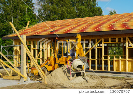 Excavator and cement mixer at wooden frame house construction site with roof beams 130070707