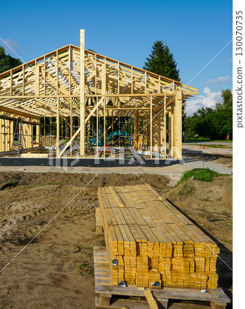 Wooden house frame under construction with stacked timber planks at building site Wooden house frame under construction with stacked timber planks at building site 130070735