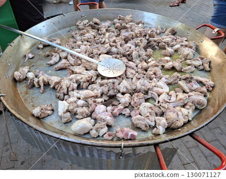A large frying pan is filled with frying chicken and liver for paella, set outside with people nearby enjoying the cooking process during a sunny day. A large frying pan is filled with frying chicken and liver for paella, set outside with people nearby enjoying the cooking process during a sunny day. 130071127