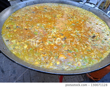 A vibrant outdoor cooking event features a large frying pan filled with colorful rice, meat, vegetables, and spices in a savory broth. The steam rises as the delicious dish simmers under the sun. 130071128