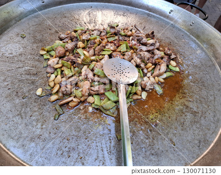 Outdoor cooking involves frying chicken and green beans in a large pan, creating a flavorful paella. The process showcases traditional cooking methods under sunny skies. 130071130