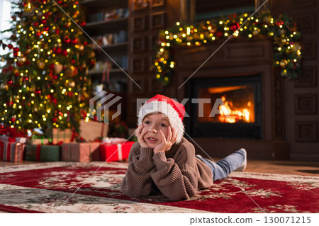 Merry Christmas. Little boy smiling near Christmas tree in classical dark interior. Young happy kid wearing Santa Hat in living room with fireplace Christmas tree gift boxes. Christmas eve at home Merry Christmas. Little boy smiling near Christmas tree in classical dark interior. Young happy kid wearing Santa Hat in living room with fireplace Christmas tree gift boxes. Christmas eve at home 130071215
