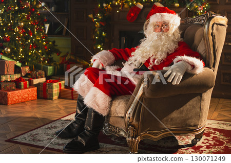 Merry Christmas. Happy Santa Claus sitting in chair in festive living room with fireplace decorations Christmas tree. Santa in cozy home on Christmas eve. Winter New Year Holiday Celebrations 130071249