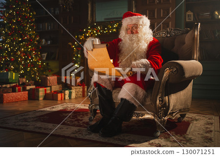 Merry Christmas. Happy Santa Claus reading wish list paper roll in festive living room with fireplace and Christmas tree. Santa with xmas wishlist letter on Christmas eve. Winter Holiday Celebrations 130071251