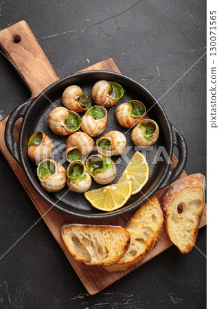 Snails escargots with garlic butter and lemon, served in a black pan with toasted bread baguette slices on black rustic background 130071565