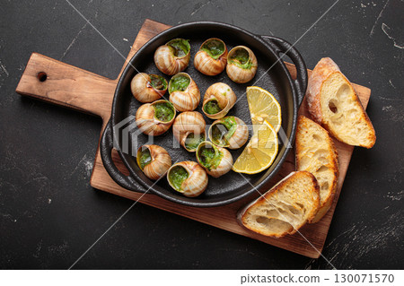 Snails escargots with garlic butter and lemon, served in a black pan with toasted bread baguette slices on black rustic background Snails escargots with garlic butter and lemon, served in a black pan with toasted bread baguette slices on black rustic background 130071570