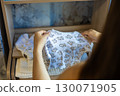 Pregnant woman holding soft baby clothes featuring a charming teddy bear pattern, organizing the nursery with love and tenderness while preparing for the joyful arrival of her newborn. 130071905