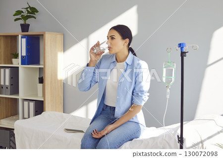 Woman drinking water while receiving vitamin solution infusion through intravenous drip 130072098