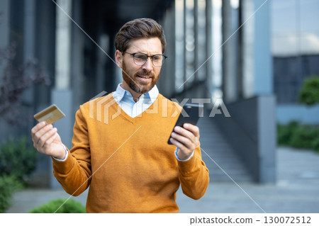 A man wearing glasses and an orange sweater holds a phone and credit card, looking concerned. Photographed outdoors conveying feelings of frustration, payment issues, or digital error. 130072512