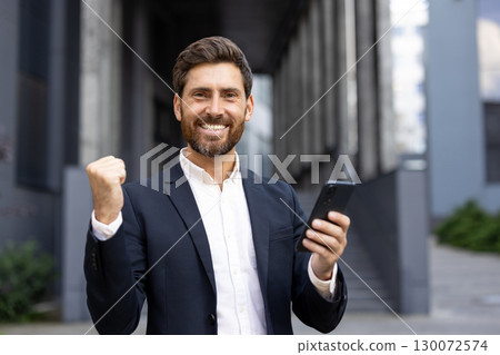 A confident businessman in a suit expressing happiness and success while holding a phone. Celebratory pose signifies achievement and satisfaction in urban surroundings. A confident businessman in a suit expressing happiness and success while holding a phone. Celebratory pose signifies achievement and satisfaction in urban surroundings. 130072574