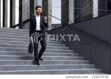 Businessman wearing a formal suit using his smartphone, walking, and carrying a bag outdoors, showcasing professionalism, connectivity, and a vibrant urban environment 130072576