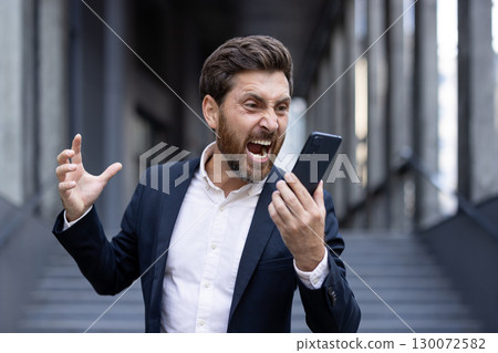 A professionally dressed man visibly shouting in frustration at his smartphone in an urban environment. His expression conveys deep anger or annoyance 130072582
