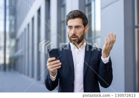 A professional man in a suit expressing shock while looking at his phone. The background showcases modern architecture, emphasizing the urban setting and suggesting a concept of unexpected news A professional man in a suit expressing shock while looking at his phone. The background showcases modern architecture, emphasizing the urban setting and suggesting a concept of unexpected news 130072613