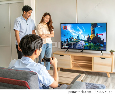 一名中學生在不感興趣的家人面前，興致勃勃地在大螢幕電視上玩線上電玩遊戲 130073052