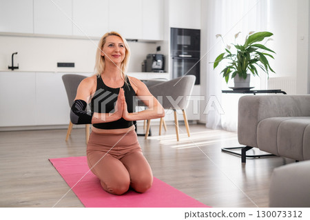 Blonde middle aged woman in sportswear standing on yoga mat in namaste pose with hands together at chest, focusing on balance and mindfulness in modern living room 130073312