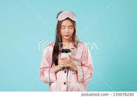 Tired woman with sleep mask holds coffee, walking up in pink pajama, relaxing, holding her paper cup, young and drinking coffee this morning, headshots isolated studio blue background Tired woman with sleep mask holds coffee, walking up in pink pajama, relaxing, holding her paper cup, young and drinking coffee this morning, headshots isolated studio blue background 130076280