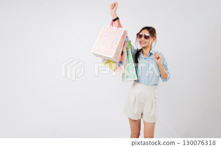 Happy young Asian woman shopping, raising hand, carrying paper bags after Black Friday, she excited and having fun dancing with a winner gesture holding her shopping bags, isolated white background 130076318