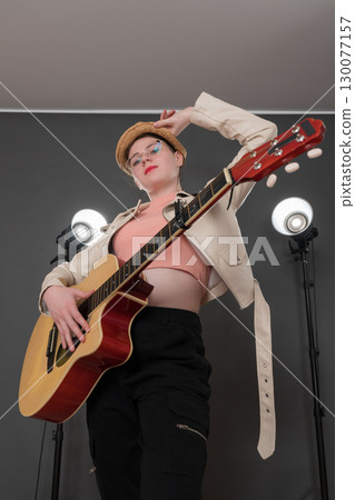 Portrait of confident female guitarist holding guitar, looking at camera. With one hand she holds brim of her straw hat, her stage outfit complemented by leather jacket and jeans. Low angle view 130077157
