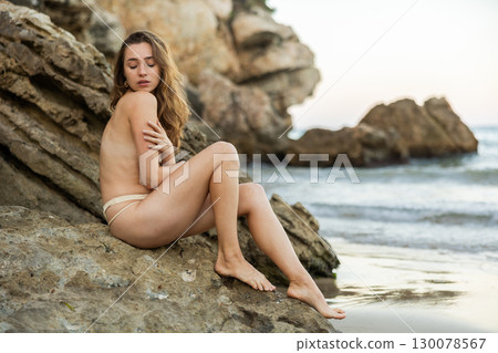 Topless girl in panties posing near sea, model sitting on large boulder Topless girl in panties posing near sea, model sitting on large boulder 130078567