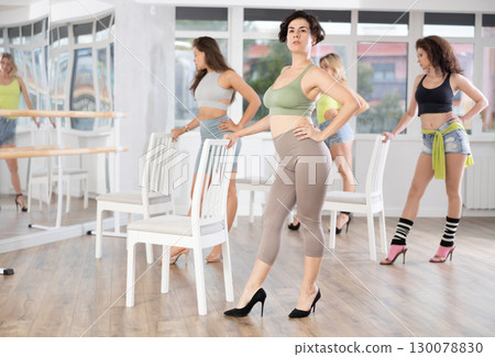 Active female dancer with group of women of different ages practicing strip plastic movements near chair in training hall during dancing classes 130078830